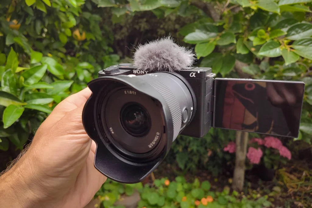 Sony ZV-E10 II зі світлосильним фіксом Sigma 11mm f/1.8 DC DN у руці, поворотний екран розгорнуто вбік, на тлі зелень саду